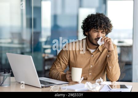 Un giovane indiano malato lavora in un ufficio. Seduto a tavola, non si sente bene, tenendo una tazza di bevanda e medicine, pulendogli il naso con un tovagliolo da un naso che cola. Foto Stock