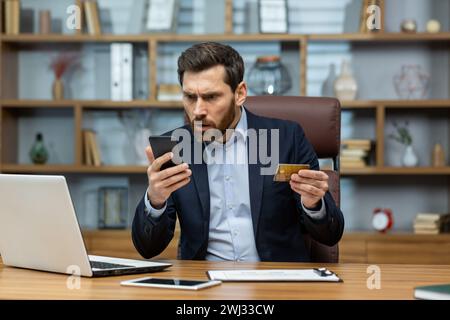 Un giovane uomo d'affari preoccupato che avesse problemi con la carta di credito. Stare seduti in ufficio alla scrivania e guardare in modo scorretto lo schermo del telefono cellulare. Foto Stock