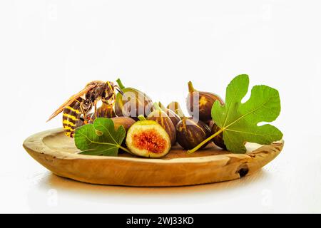 Una frusta e frutta fresca di fico cruda, una porzione tagliata e una foglia di fico su un tavolo, su un piatto di legno isolato Foto Stock