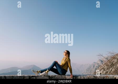 La giovane donna siede su un recinto di pietra tra le montagne appoggiate alla sua mano. Vista laterale Foto Stock