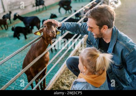 La bambina si trova accanto a suo padre che accarezza una testa di capra che sbuca su una recinzione Foto Stock