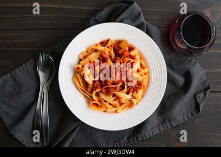Ciotola di spaghetti di pappardelle condita in autentica salsa bolognese: Primo piano di una ciotola di spaghetti poco profonda con un bicchiere di vino rosso, una forchetta e un cucchiaio Foto Stock