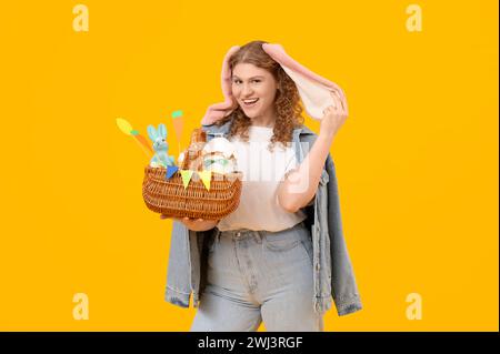 Bella giovane donna felice con orecchie conigliette con cesto di Pasqua su sfondo giallo Foto Stock