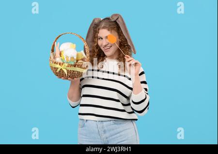 Bella giovane donna felice con orecchie conigliette con cestino di Pasqua e uovo di carta su sfondo blu Foto Stock