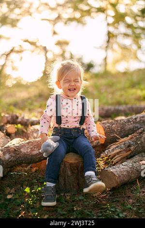 La bambina siede su un ceppo nella foresta con un giocattolo in mano e ride con gli occhi chiusi Foto Stock