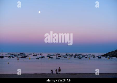 Una famiglia sulla spiaggia al tramonto con barche ancorate al largo di Tamariu, Catalogna, Spagna. Foto Stock