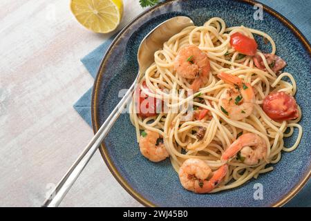 Spaghetti con gamberetti, pomodori ed erbe aromatiche in salsa di limone in un piatto blu con cucchiaio su un tavolo di legno bianco, Mediterra fatta in casa Foto Stock