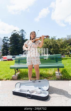 Immagine verticale di una giovane artista latina violinista che suona nel parco lavorando con la cassa per violino sul pavimento, con alberi e cielo sullo sfondo, poliziotto Foto Stock