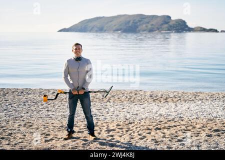 Un giovane sorridente in piedi con un metal detector in mano davanti a lui sulla spiaggia Foto Stock