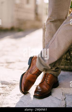 L'uomo con scarpe marroni si trova sulle pietre di pavimentazione con gambe incrociate. Ritagliato. Senza volto Foto Stock