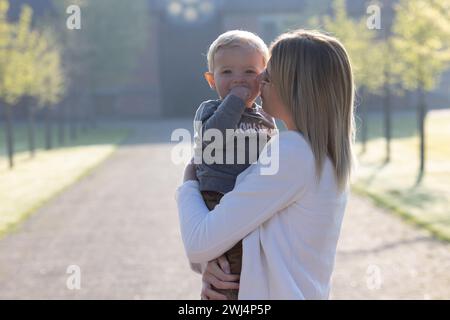 Questa immagine cattura un momento tenero tra una giovane donna caucasica e un bambino allegro. La donna tiene il bambino vicino, i volti quasi toccano, condividendo un momento di gioia e affetto. Gli occhi luminosi del bambino e il sorriso delizioso illuminano la scena tanto quanto la luce soffusa del mattino che li avvolge. Il parco intorno a loro è tranquillo e tranquillo, un ambiente perfetto per uno scambio così intimo. Momenti preziosi nella foschia mattutina. Foto di alta qualità Foto Stock