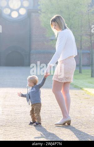 Questa scena emozionante si svolge in un parco tranquillo, dove una giovane donna caucasica tiene delicatamente la mano di un bambino, guidandolo mentre compie piccoli passi. Il sole del mattino getta una luce delicata sulla coppia, simboleggiando la speranza e l'inizio di un viaggio. La donna, vestita elegantemente con una camicetta bianca e una gonna in pizzo, guarda il bambino con una posizione protettiva, il suo linguaggio del corpo esprimendo cura e incoraggiamento. Luce guida: Il viaggio di un bambino. Foto di alta qualità Foto Stock