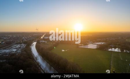 Questa fotografia aerea cattura la serena bellezza dell'alba in un ambiente rurale. Il sole del mattino presto rompe l'orizzonte, bagnando il paesaggio in una luce soffusa e diffusa. Una turbina eolica si trova in lontananza, simboleggiando una vita sostenibile in mezzo all'ambiente naturale. Un canale serpeggiante riflette la luce del mattino, creando un percorso di luminosità attraverso i campi. La comunità rurale sembra svegliarsi per una mattinata tranquilla, con il potenziale dei giorni che si stanno svolgendo prima di essa. L'alba si ferma sulla turbina eolica e sul canale in campagna. Foto di alta qualità Foto Stock