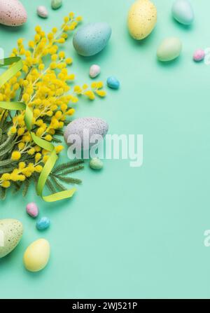 Una disposizione creativa di uova pasquali color pastello, molle mimosa e spolverini di caramelle su una superficie verde morbida, simboleggiando i festeggiamenti primaverili Foto Stock