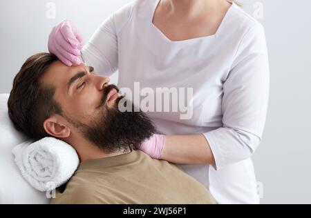 Primo piano dell'uomo bearded che ottiene l'iniezione di bellezza alla clinica estetica. Medico che inietta il riempitivo anti-invecchiamento nel bel viso maschile Foto Stock