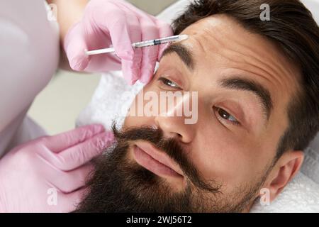 Primo piano dell'uomo bearded che ottiene l'iniezione di bellezza alla clinica estetica. Medico che inietta il riempitivo anti-invecchiamento nel bel viso maschile Foto Stock