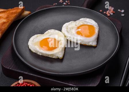 Due uova fritte a forma di cuore su un piatto di ceramica nera su uno sfondo di cemento scuro Foto Stock