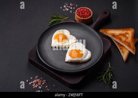 Due uova fritte a forma di cuore su un piatto di ceramica nera su uno sfondo di cemento scuro Foto Stock