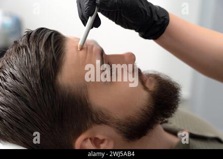 Ritratto di un giovane e bello uomo con la fronte rugosa. le mani di un cosmetologo tengono in mano una matita cosmetica e apportano segni sul Foto Stock