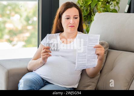 Foto di una donna incinta confusa che legge un opuscolo prima di prendere una pillola a casa Foto Stock