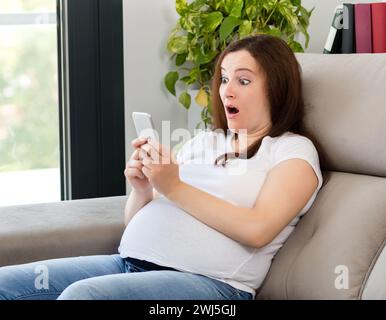 Stupita donna incinta che guarda uno smartphone a casa Foto Stock