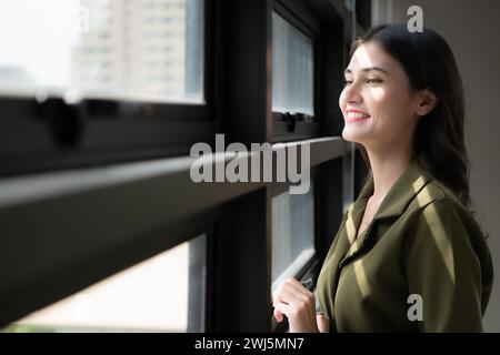 Ritratto di una bella giovane donna in piedi vicino alla finestra e sorridente Foto Stock