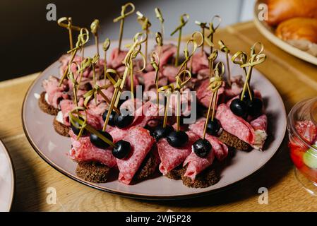 Eleganti involtini di salame con formaggio spalmabile e olive nere, serviti come antipasti con pane di segale su un piatto di ceramica, ideali per feste e riunioni. Foto Stock