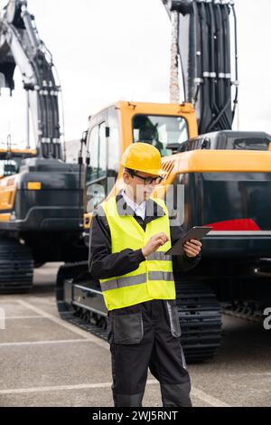 Ingegnere in un casco con un tablet digitale stand accanto agli escavatori da cantiere. Foto Stock