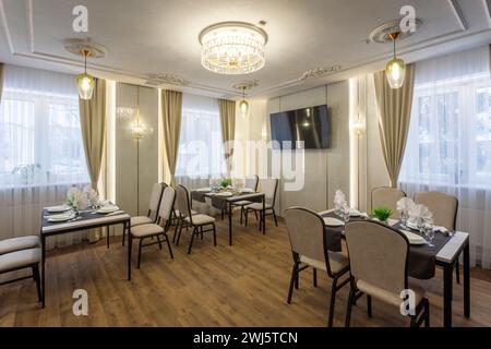 Interni moderni di una sala banchetti con posti da pranzo. Morbide sedie beige attorno ai tavoli servite con bicchieri, piatti e tovaglioli in interni classici. Foto Stock