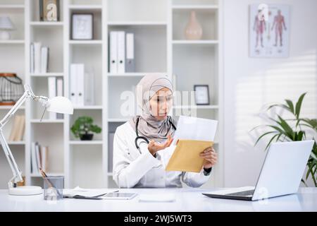 Una donna musulmana concentrata in un hijab e cappotto bianco esamina i documenti nel suo studio medico, simboleggiando la diversità sanitaria. Foto Stock