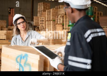 Giovani operai di magazzino di sesso maschile e femminile che utilizzano tablet digitali per lavorare in una fabbrica di legno, controllando la correttezza delle tavole Foto Stock
