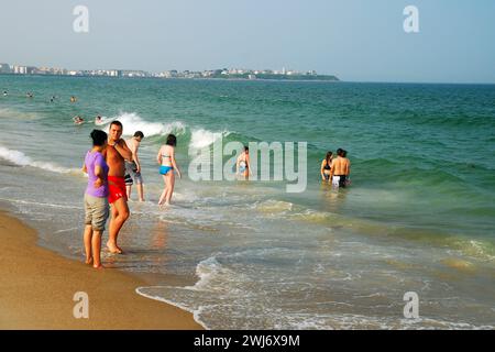 Una coppia di giovani adulti si tuffa nelle acque della spiaggia mentre altri giocano tra le onde in una giornata di vacanza estiva Foto Stock