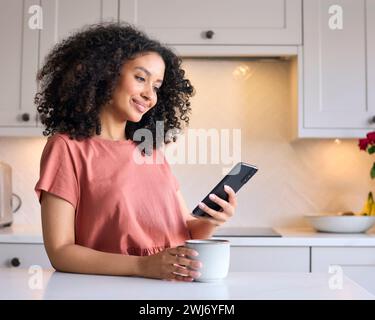 Giovane donna appoggiata al bancone della cucina a casa che controlla i social media sul telefono cellulare con bevanda calda Foto Stock