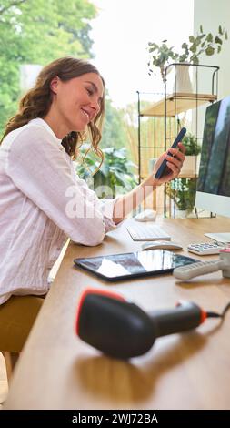 Donna che gestisce un business online da casa e lavora al computer utilizzando il telefono cellulare Foto Stock