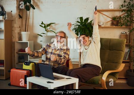 Coppia senior, uomo e donna anziani che festeggiano con successo la prenotazione del prossimo viaggio, seduti con i bagagli pieni Foto Stock