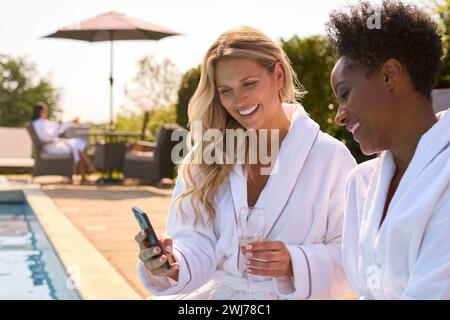 Due amiche mature con telefono cellulare in accappatoio all'aperto in piscina e champagne il giorno della Spa Foto Stock