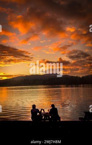 Due persone sorseggiano una bevanda tranquilla che si staglia sul tramonto sulla riva del lago Windermere. Foto Stock