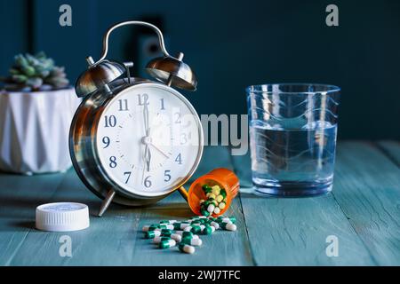 Apri una bottiglia di pillole prescritte accanto a una sveglia e un bicchiere d'acqua sul comodino in legno. Messa a fuoco selettiva con sfondo sfocato. Foto Stock