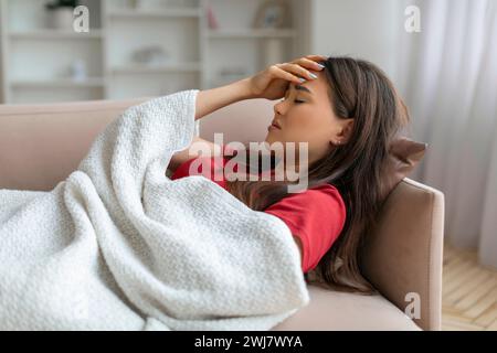 Donna asiatica esausta che tocca la fronte, sdraiata sul divano sotto la coperta Foto Stock