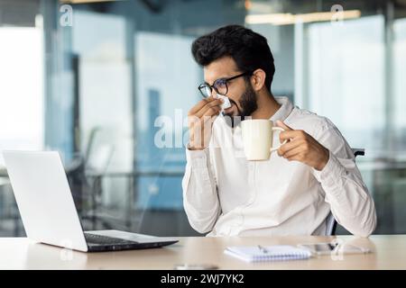 Giovane uomo d'affari indiano malato, lavoratore d'ufficio seduto alla scrivania usando il computer portatile, tenendo una tazza di bevanda e asciugando il naso con un fazzoletto dal naso che cola. Foto Stock