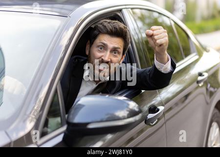 Un giovane arrabbiato e irritato con un costume da lavoro guarda fuori dal finestrino della macchina. È al volante, grida alla telecamera e agita le mani minacciosamente. Foto in primo piano. Foto Stock