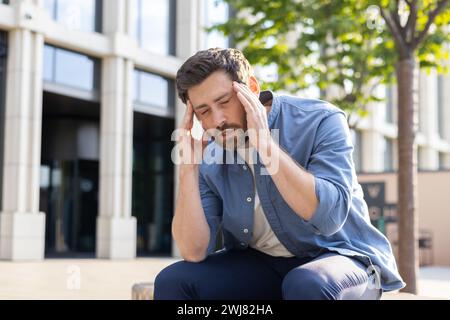 Un uomo d'affari maturo in difficoltà che sfrega i templi a causa del mal di testa, seduto accanto all'edificio degli uffici, sentendo lo stress del lavoro e il burnout. Foto Stock