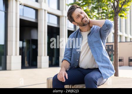 Un uomo d'affari maturo con la barba sembra scomodo con il dolore al collo mentre siede fuori da un ufficio, mostrando un problema comune sul posto di lavoro. Foto Stock