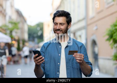Uomo sorridente con la barba in un abbigliamento casual che utilizza uno smartphone e tiene in mano una carta di credito in una vivace strada della città la sera. Foto Stock