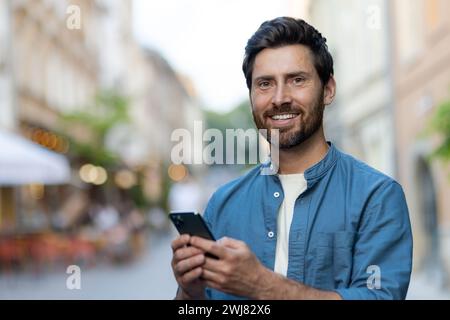 Allegro uomo barbuto maturo che utilizza lo smartphone in un ambiente urbano vivace al tramonto, trasudando fiducia in un ambiente informale. Foto Stock