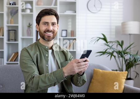 Allegro uomo maturo con barba che si rilassa a casa, comodamente seduto su un divano, allegramente con uno smartphone. Foto Stock