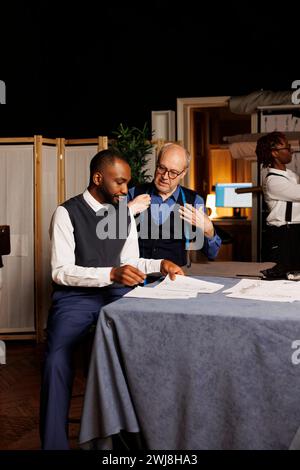 Un cliente elegante che parla delle opzioni di personalizzazione della tuta migliore con un esperto preparatore di valigie sugli schizzi di abbigliamento in un negozio di sartoria. Gentiluomo che guarda i disegni di moda, sceglie le regolazioni desiderate Foto Stock