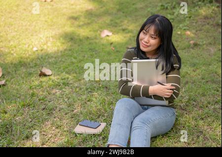 Una bella studentessa asiatica del college è seduta sull'erba in un parco universitario con il suo portatile. Foto Stock