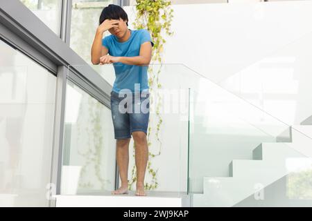 Un ragazzo asiatico adolescente sta in piedi su una scala moderna a casa, sembra stressato e triste Foto Stock