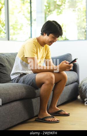 Adolescente asiatico si siede su un divano a casa, invischiato nel suo smartphone Foto Stock